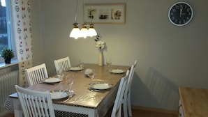 Uma sala de jantar com uma mesa posta para quatro pessoas, cadeiras brancas, um vaso com flores e um relógio na parede.