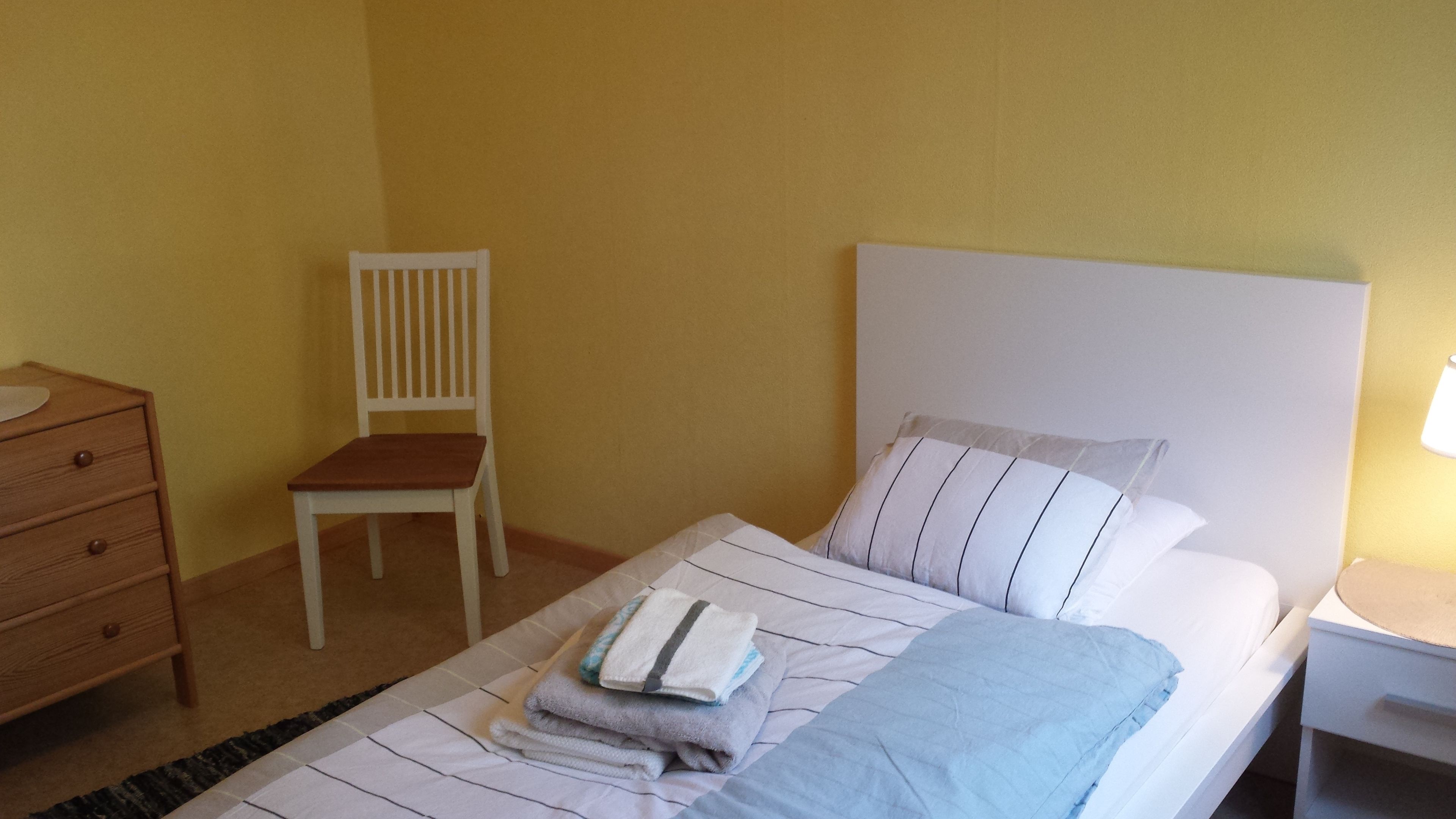 Una cama individual con cabecera blanca, una silla de madera, una mesita de noche con lámpara y un aparador de madera con espejo.