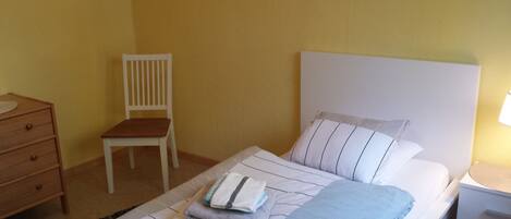 Una cama individual con cabecera blanca, una silla de madera, una mesita de noche con lámpara y un aparador de madera con espejo.