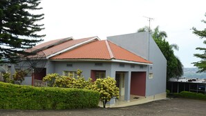 A two-story building with a red-tiled roof, a grey facade, and a small front yard with trimmed hedges and flowering plants. - Tinho Guest House (Kigali)