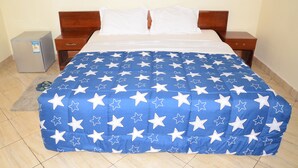 A bedroom with a bed covered in a blue and white star-patterned blanket, wooden headboard, white pillows, two bedside tables, and a small gray refrigerator. - Tinho Guest House (Kigali)