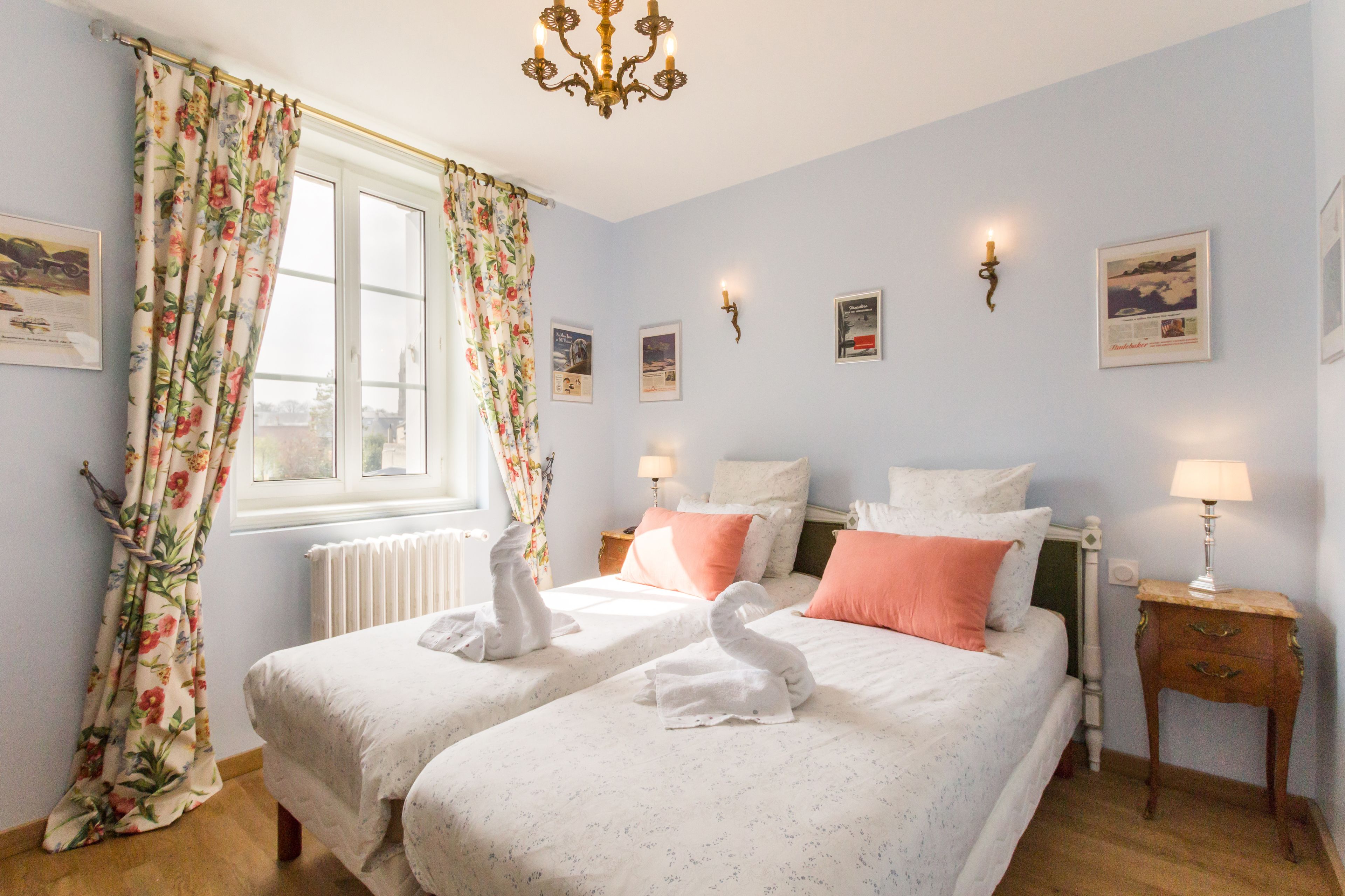 A bedroom with two beds, floral curtains, a chandelier, wall-mounted lamps, and framed pictures.