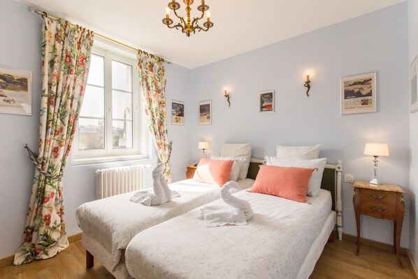 A bedroom with two beds, floral curtains, a chandelier, wall-mounted lamps, and framed pictures. - DDAY Aviators Le Manoir (Arromanches-les-Bains)