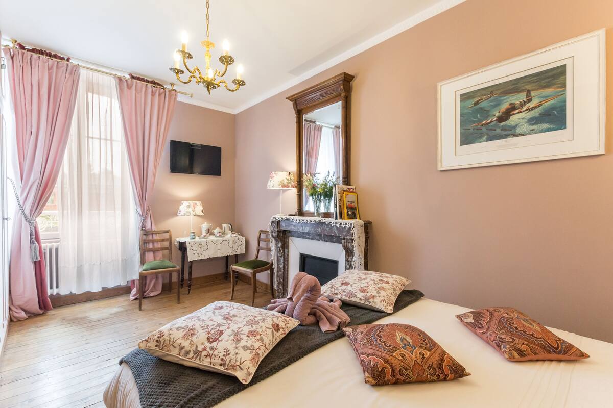 Un dormitorio con cama, almohadas, una silla, un escritorio, chimenea y un cuadro en la pared.