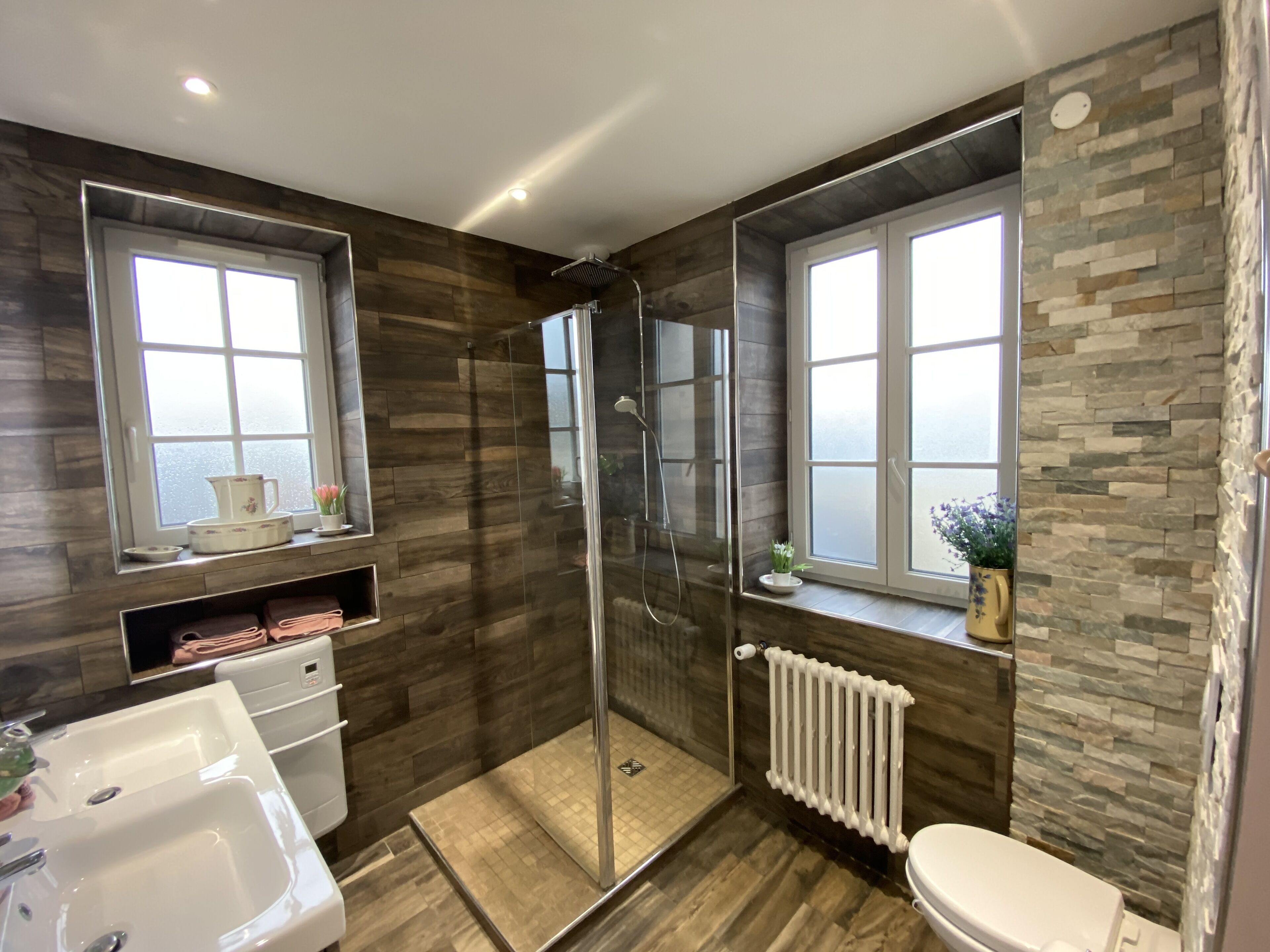A modern bathroom with a shower, toilet, and sink. There are windows with views and a shelf with toiletries.