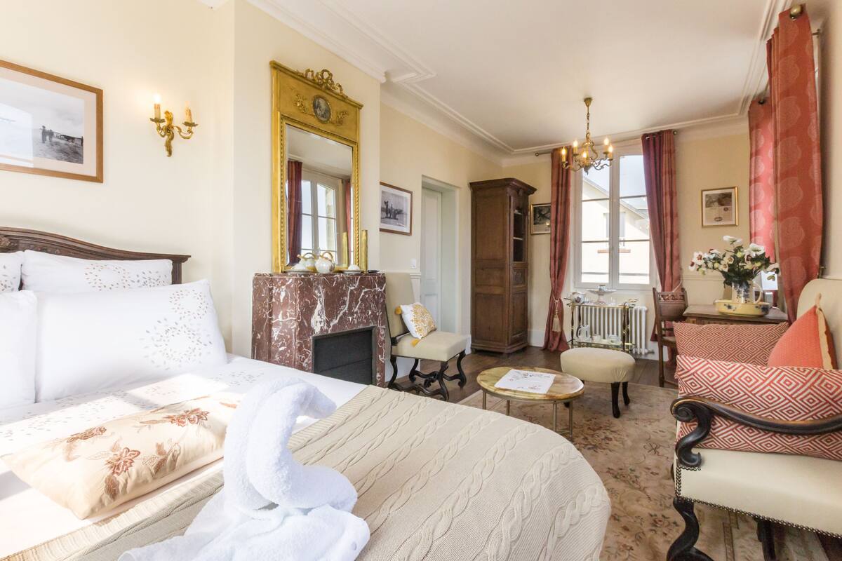 Habitación de hotel con una cama grande, una zona de estar con una silla y una mesa, una chimenea y vistas al exterior.
