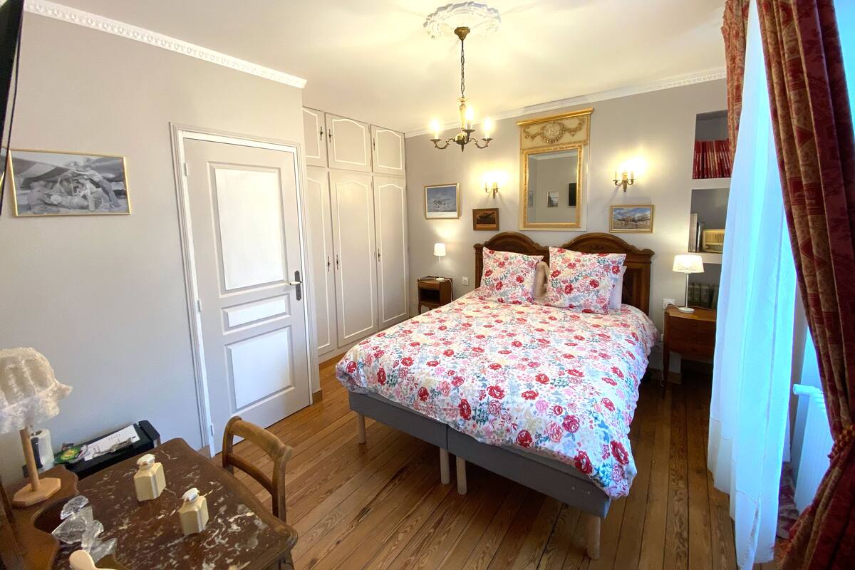 Un dormitorio con una cama, suelo de madera, paredes blancas, una araña y un espejo, además de una puerta.