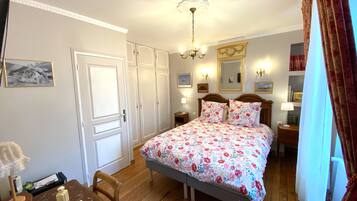 Un dormitorio con una cama, suelo de madera, paredes blancas, una araña y un espejo, además de una puerta.