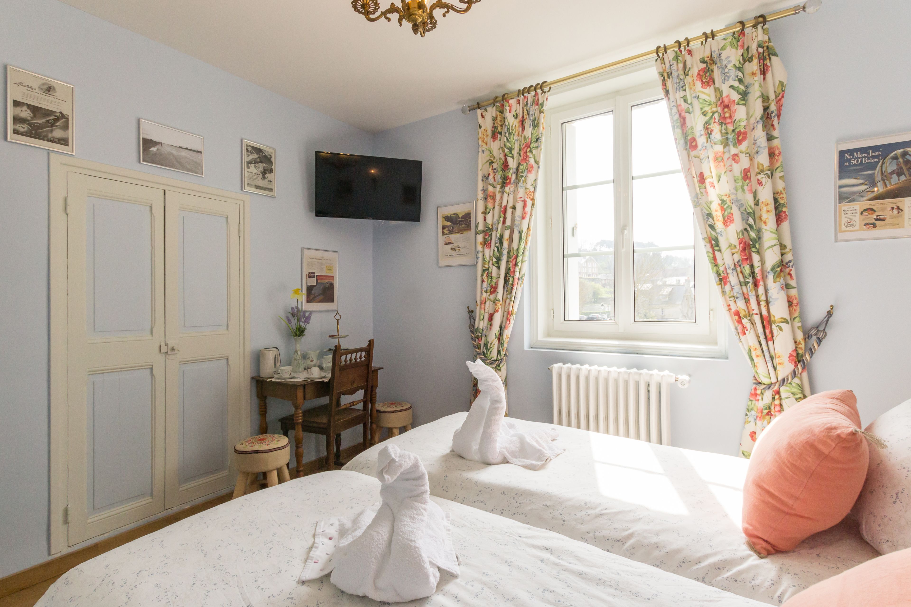 A bedroom with two beds, a television, a desk, and a window with floral curtains.