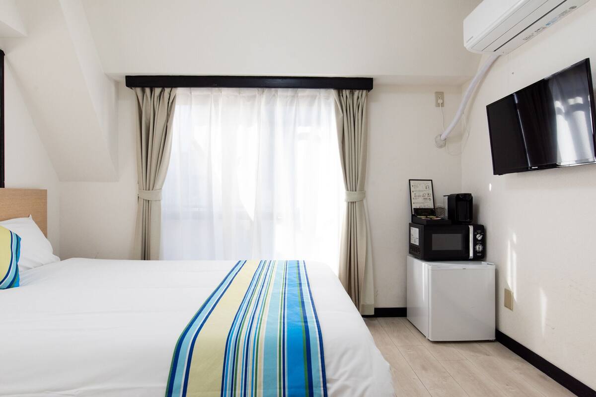 A hotel room with a bed, a small kitchenette, a TV, and a window with curtains.