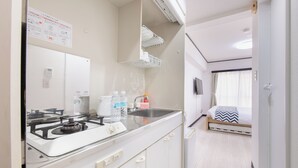 A compact kitchenette with a stove, sink, and storage cabinets. A bed with a striped blanket is visible in the adjacent room. - G-stays Shinjuku (Tokyo)