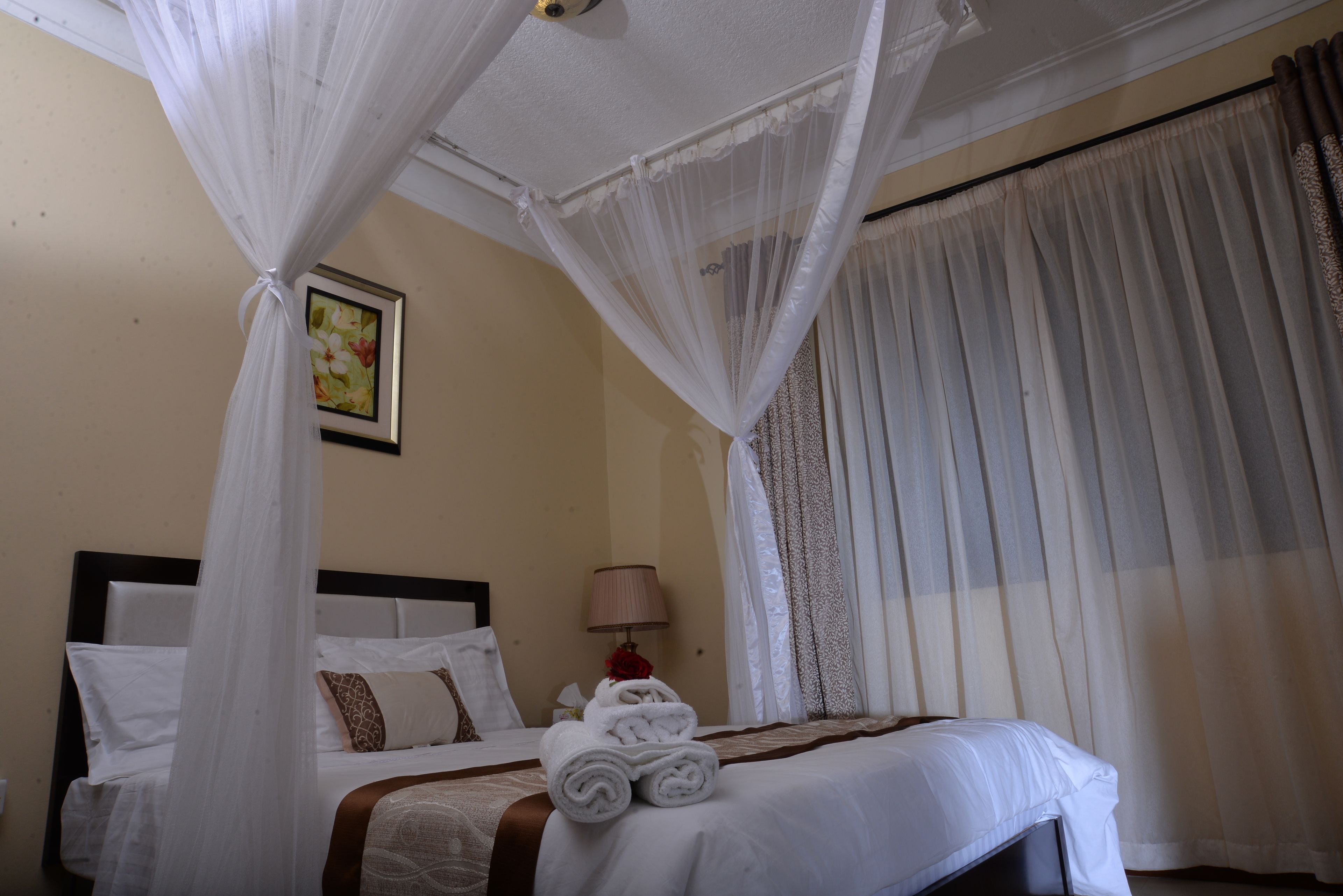 A hotel room with a bed, white curtains, a bedside lamp, and a framed picture on the wall.