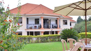 A two-story house with a balcony, surrounded by a well-maintained garden and a dining area with chairs and an umbrella. - Royal Victoria House (Kampala)
