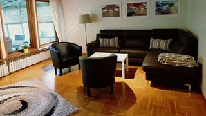 Ein Wohnzimmer mit Holzboden, einem schwarzen Ledersofa, einem weißen Couchtisch und einem schwarzen Sessel. An der Wand hängen drei gerahmte Bilder, eine Stehlampe und ein Fenster mit Vorhängen.
