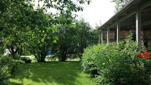 Ein gepflegter Garten mit verschiedenen Bäumen und Sträuchern, einer Rasenfläche und einem Gebäude mit überdachter Veranda.