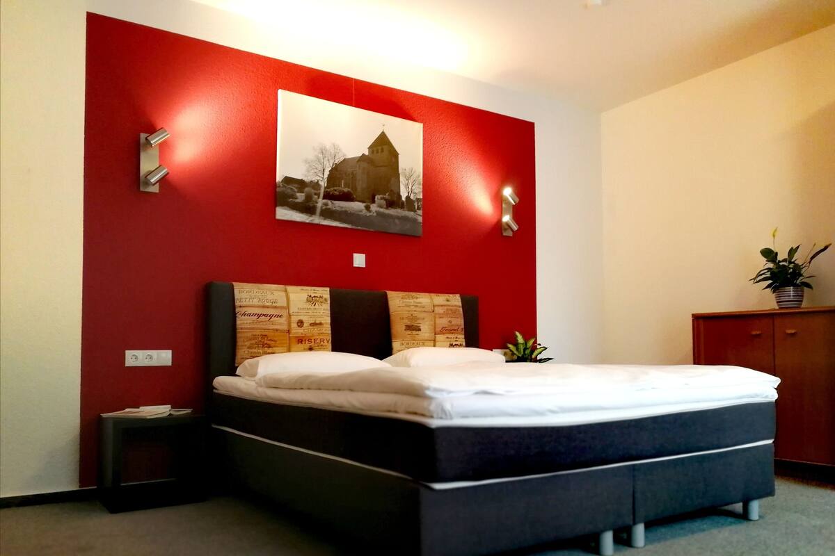 A bedroom with a bed, a red accent wall, a framed picture, a nightstand, and a small plant.
