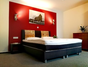 A bedroom with a bed, a red accent wall, a framed picture, a nightstand, and a small plant.