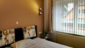 A hotel room with a bed, a potted plant, a window with curtains, and a wall-mounted light fixture. - Hotel & Gasthaus Zum Domkreuger (Walsrode)