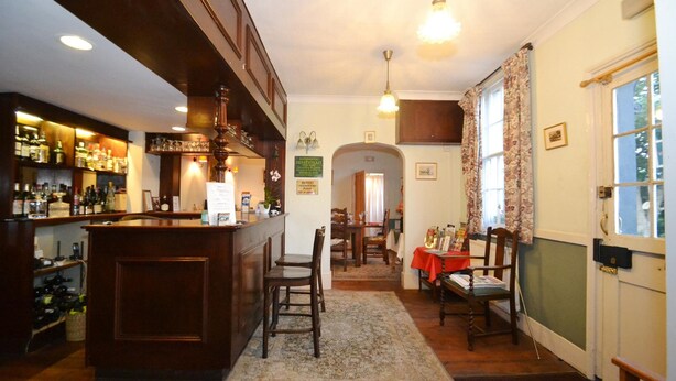 Uma área de bar com balcão de madeira, várias garrafas e copos. Há também uma sala de jantar com uma mesa e cadeiras, e um tapete no chão.