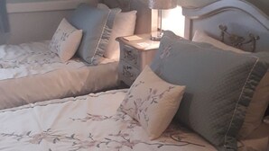 Uma cama de casal com roupa de cama florida e várias almofadas, uma mesa de cabeceira com abajur e um criado-mudo com telefone.