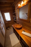 Une salle de bain rustique, avec une cabine en bois, un lavabo en bois, un miroir et une fenêtre équipée de stores.