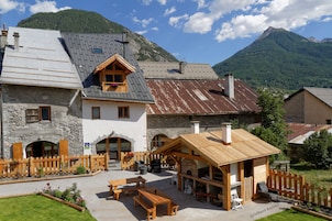 Un chalet de montagne rustique, avec des murs en pierre, des bancs en bois et un espace extérieur aménagé avec des sièges sous un auvent.
