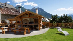 Un'area pranzo esterna rustica con panche di legno e una griglia per barbecue, circondata da un recinto in legno e un prato.