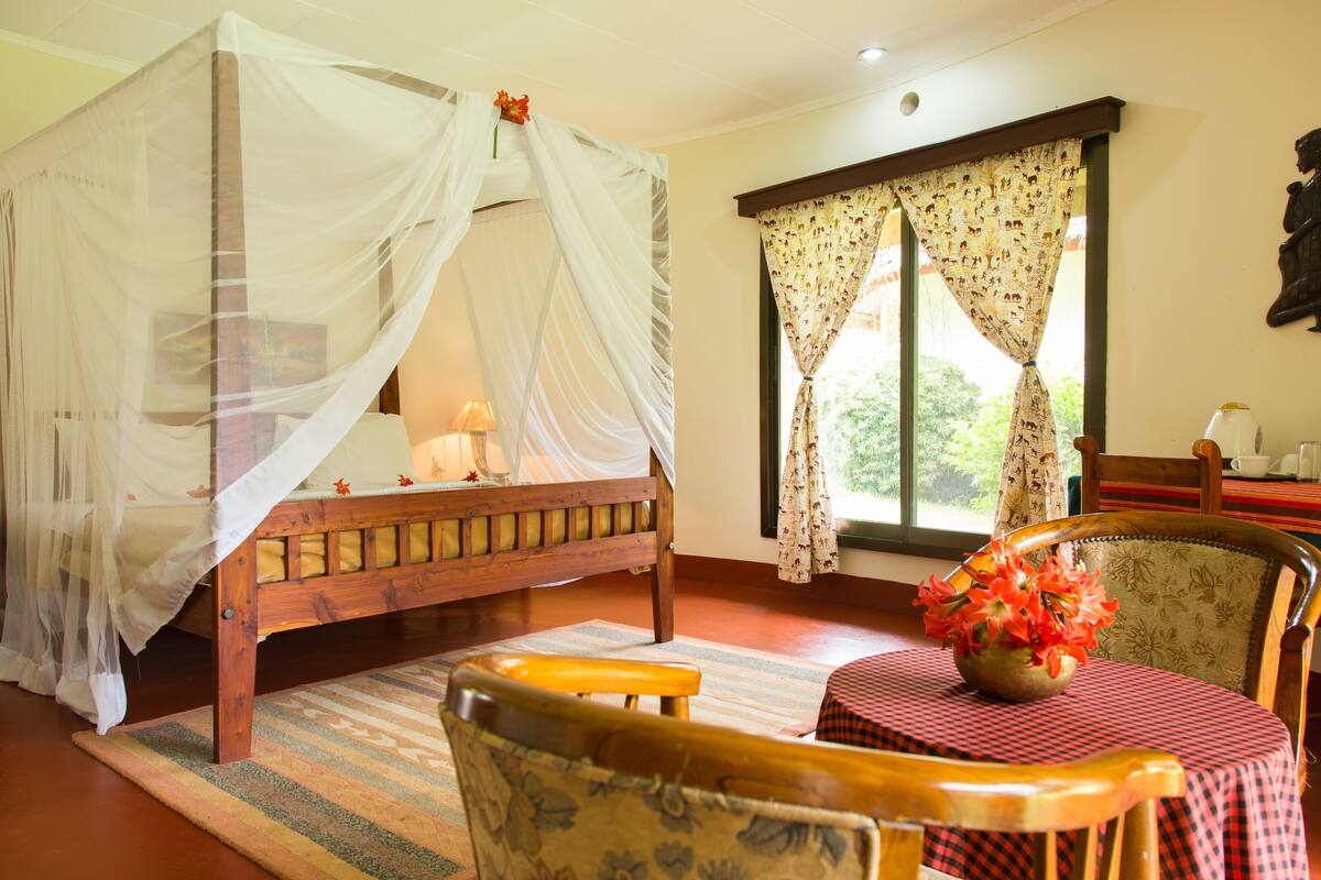 A room with a four-poster bed, a wooden chair, a small table with a vase of flowers, and a window with curtains.