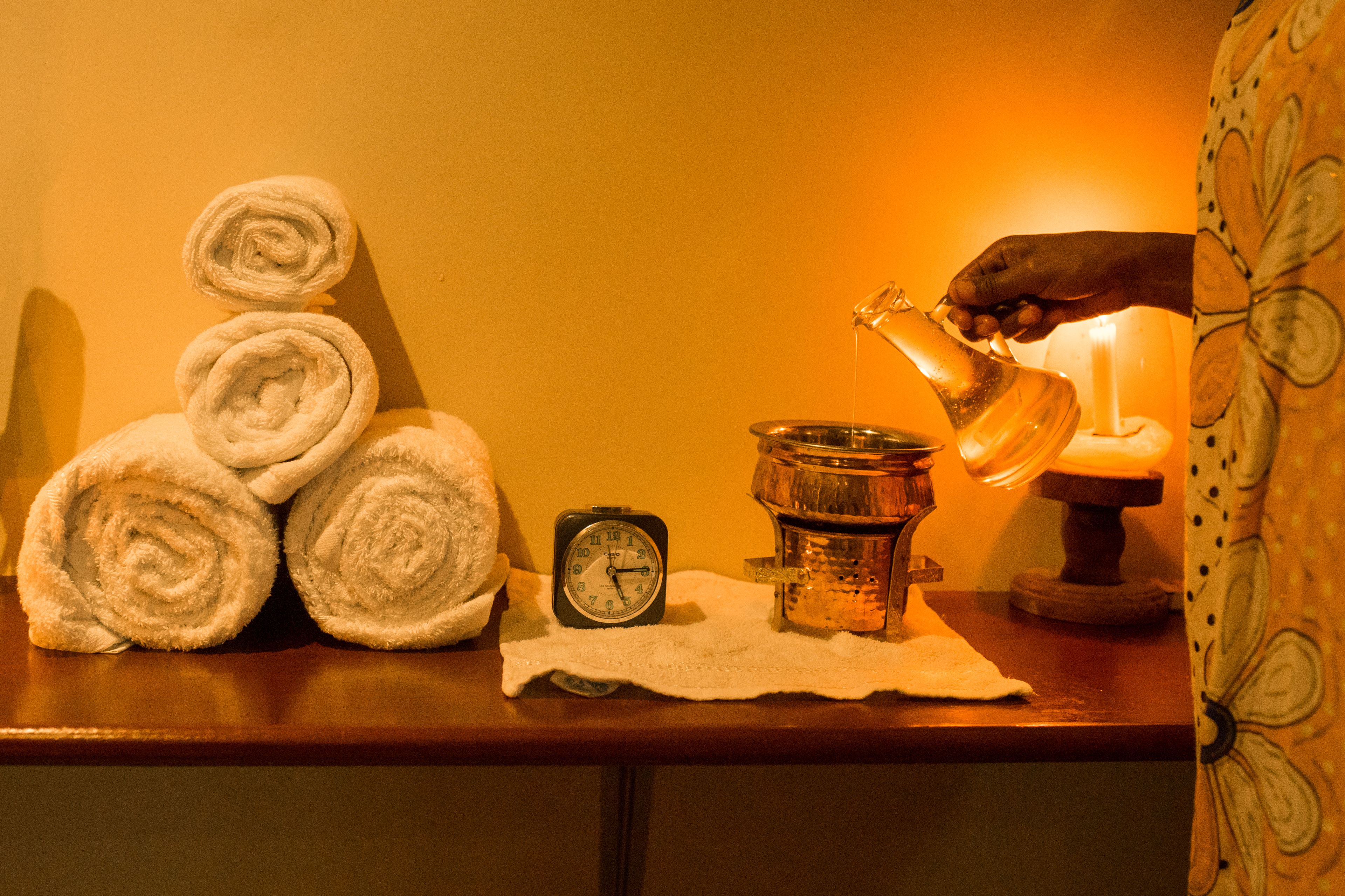 A wooden table with rolled towels, a clock, a candle, and a hand pouring water from a jug.
