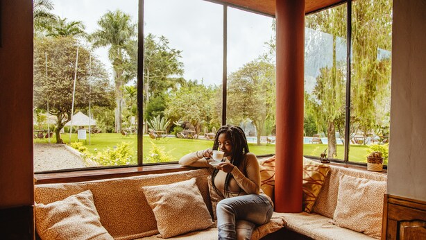 A woman sitting on a sofa by a large window with a view of a garden. - Kilemakyaro Mountain Lodge (Moshi)