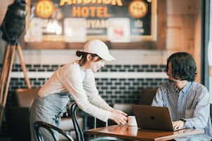 Una persona con gorra blanca y pantalones a rayas de pie junto a una mesa con una computadora portátil y una taza blanca, en una habitación con un letrero que dice "Anchor Hotel".