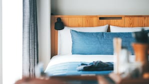 Habitación de hotel con cabecera de madera, cama con ropa de cama blanca y azul, y mesita de noche con una lámpara y una botella.