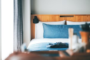 Habitación de hotel con cabecera de madera, cama con ropa de cama blanca y azul, y mesita de noche con una lámpara y una botella.