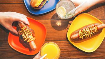 Ein Holztisch mit zwei Hotdogs auf orangefarbenen und gelben Tellern, einem Sandwich auf einem blauen Teller und einem Glas Orangensaft.