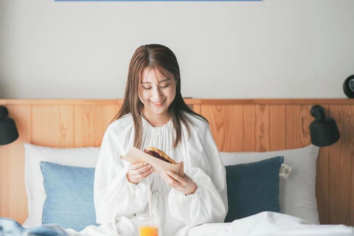 A person sitting on a bed with a wooden headboard, holding a piece of bread.
