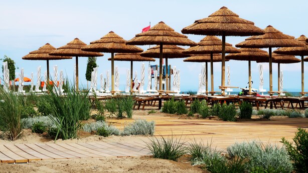 A beachside area with thatched umbrellas, wooden tables, and chairs, surrounded by greenery.