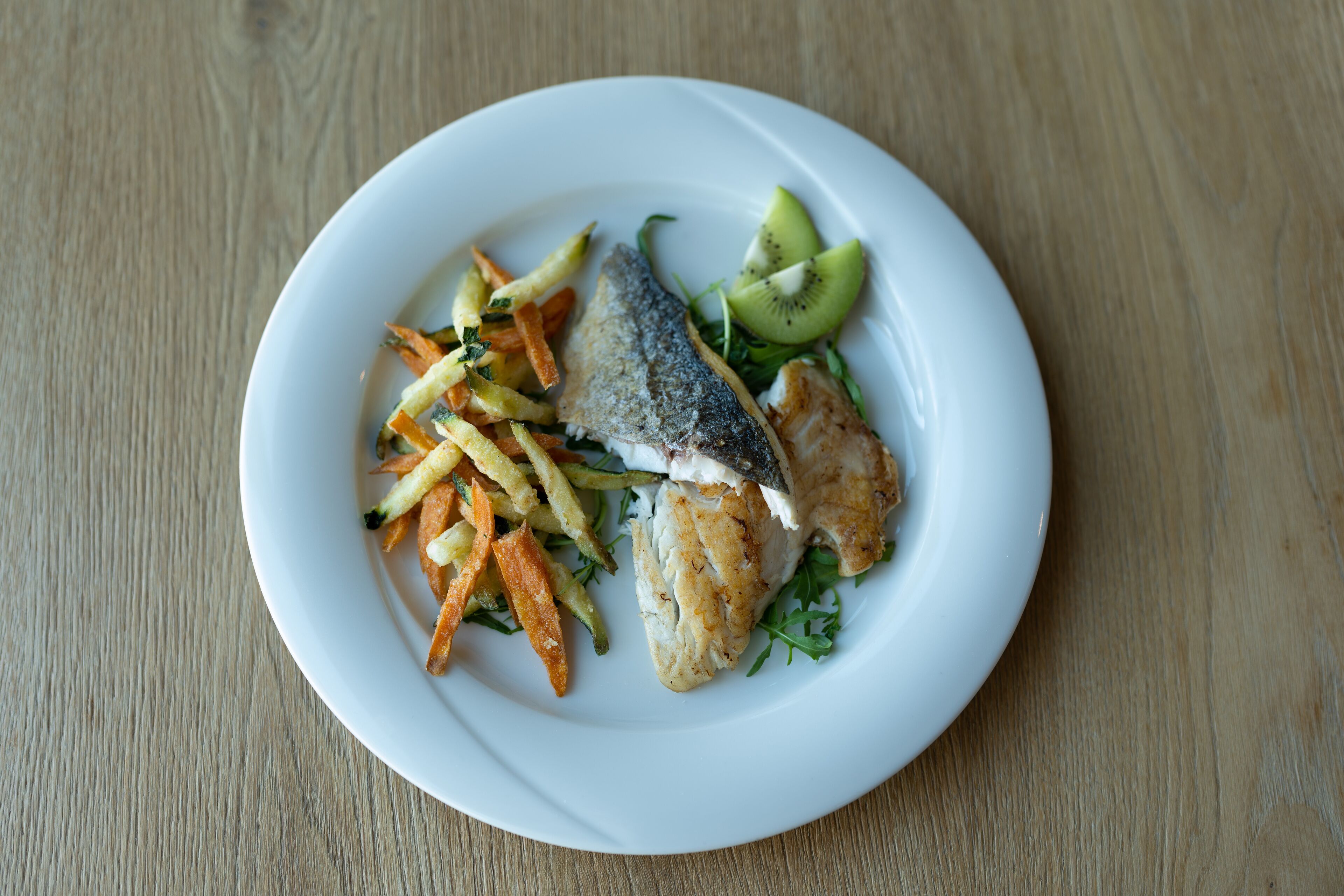 A plate with fish, fries, and a slice of kiwi on a wooden table.