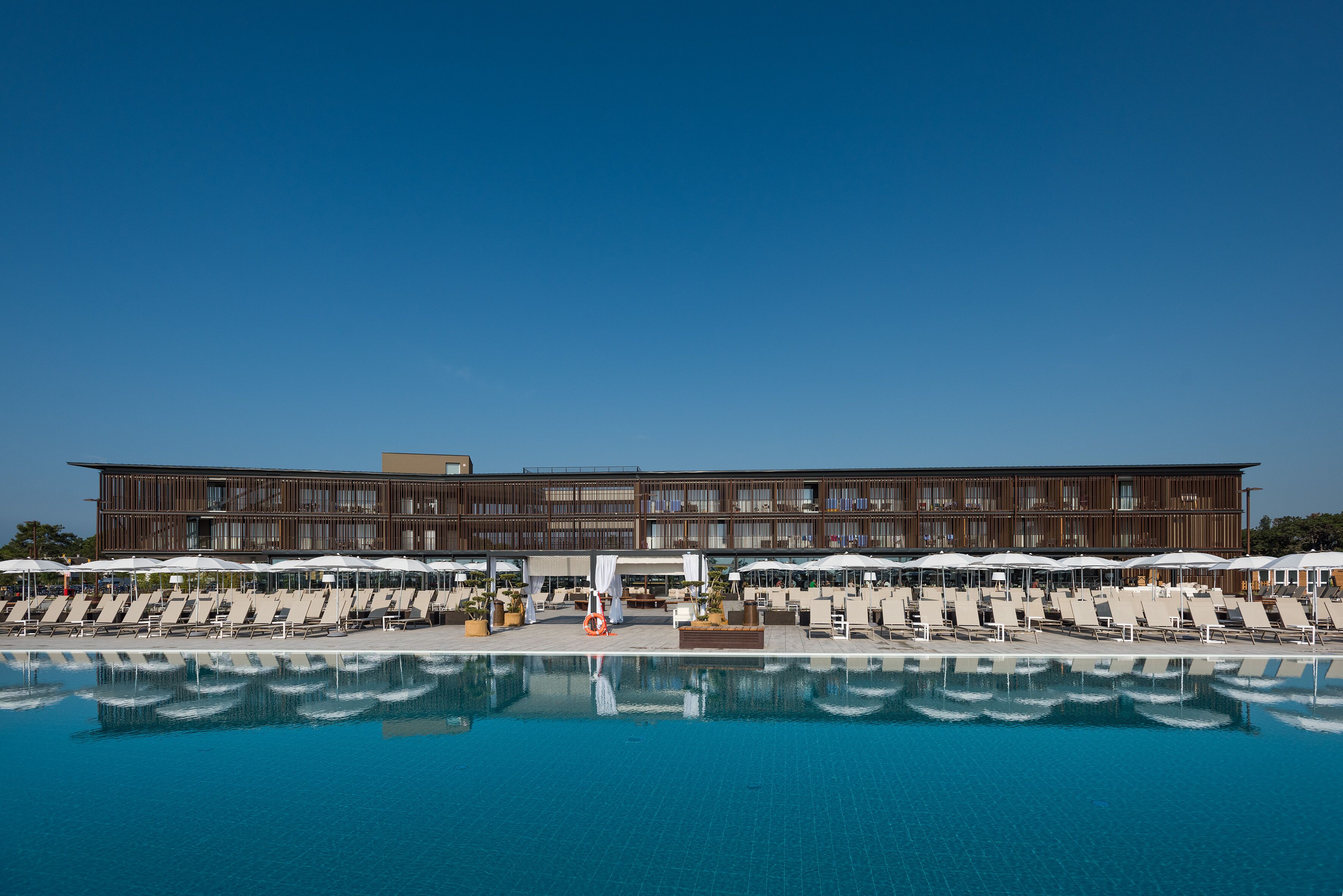 A modern hotel with a pool area, lounge chairs, umbrellas, and a clear blue sky.