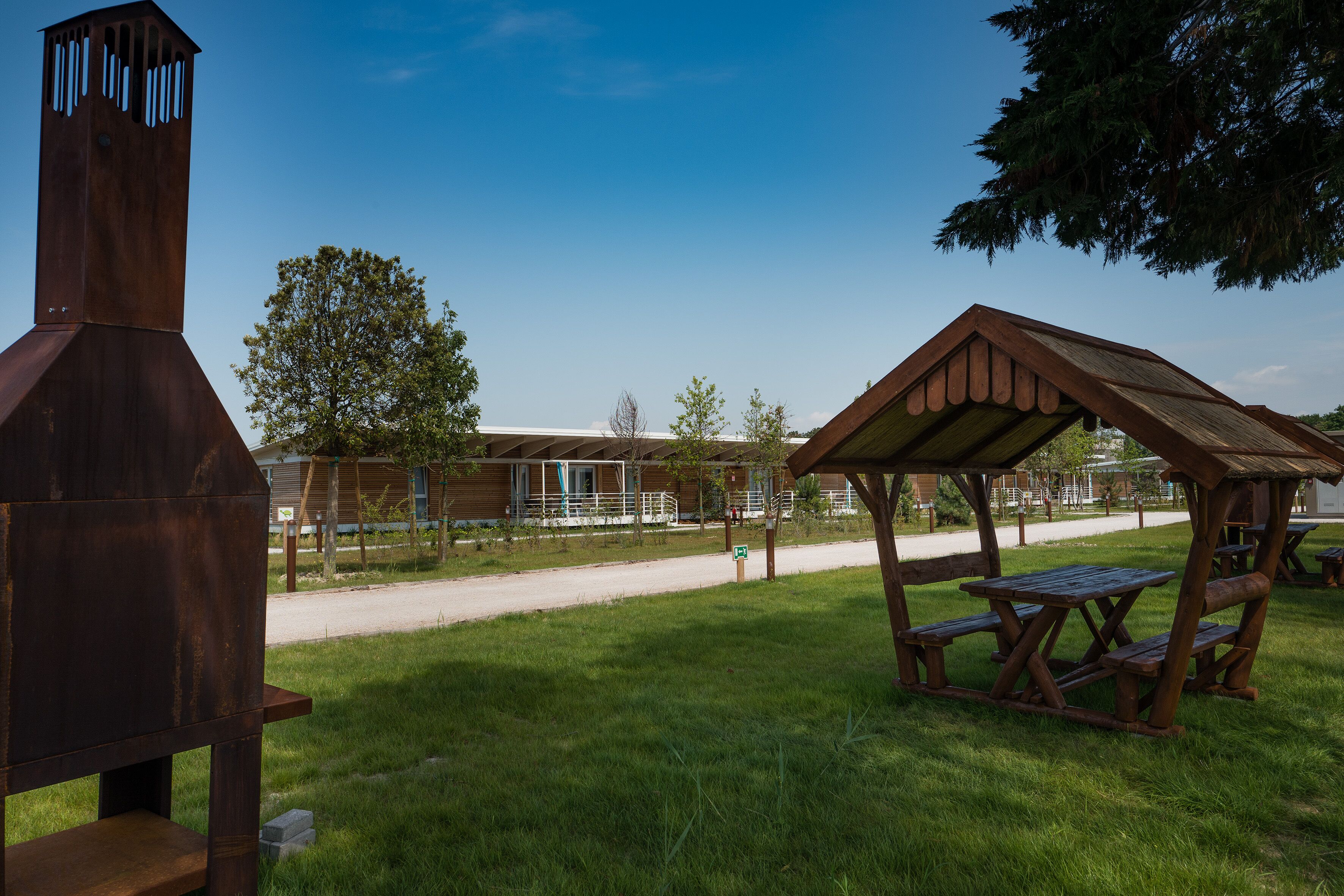 Ein rustikaler Außenbereich mit einer Holzkohlegrillstation und Picknicktischen, umgeben von einem gepflegten Rasen und einem Gebäude im Hintergrund.