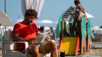 Un homme est assis sur une chaise longue sur la plage, en train de lire un journal, avec une boisson posée sur la table.