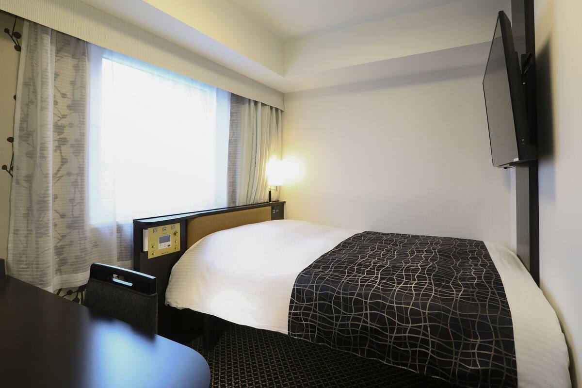 Una habitación de hotel con cama, escritorio, silla, televisor y ventana con cortinas.