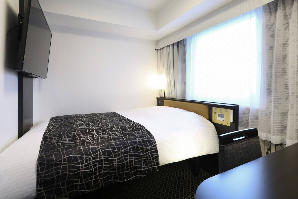 Habitación de hotel con cama, televisor montado en la pared, escritorio con silla y ventana con cortinas.