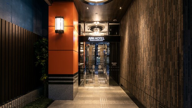 L’entrée d’un hôtel au design moderne, avec un mur en bois, une porte en verre et une enseigne indiquant « APA HOTEL ».