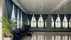 Un amplio vestíbulo con una fila de sillas negras, una planta grande en maceta y ventanas grandes con cortinas azules.