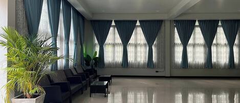 A spacious lobby with a row of black chairs, a large potted plant, and large windows with blue curtains.