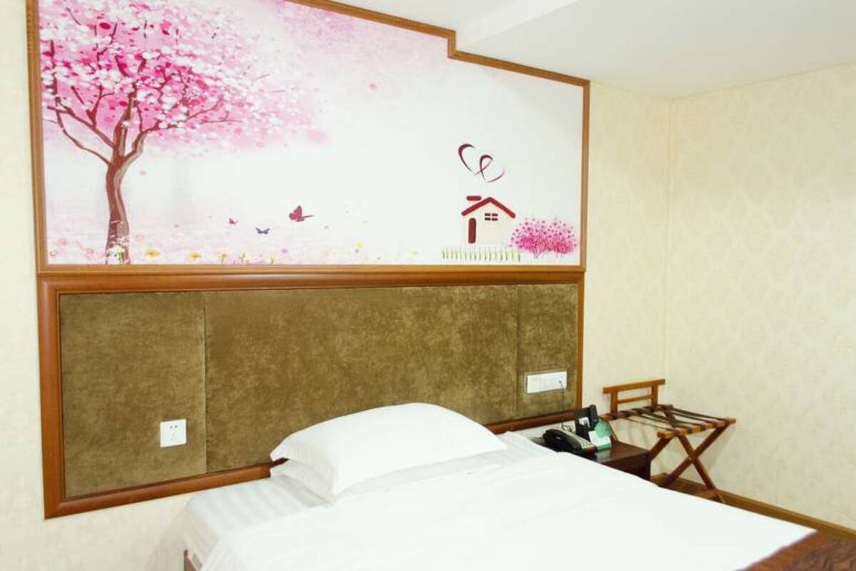 Una habitación de hotel con cama, cabecera de madera, una mesita con silla, y una pared decorada con un árbol de cerezo e ilustración de una casa.