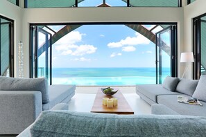 A modern living room with a large glass door leading to a balcony overlooking a beach. - Villa Moonshadow (Koh Samui)