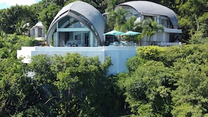 Una villa moderna con un salón envuelto en cristal y una piscina con sombrillas.