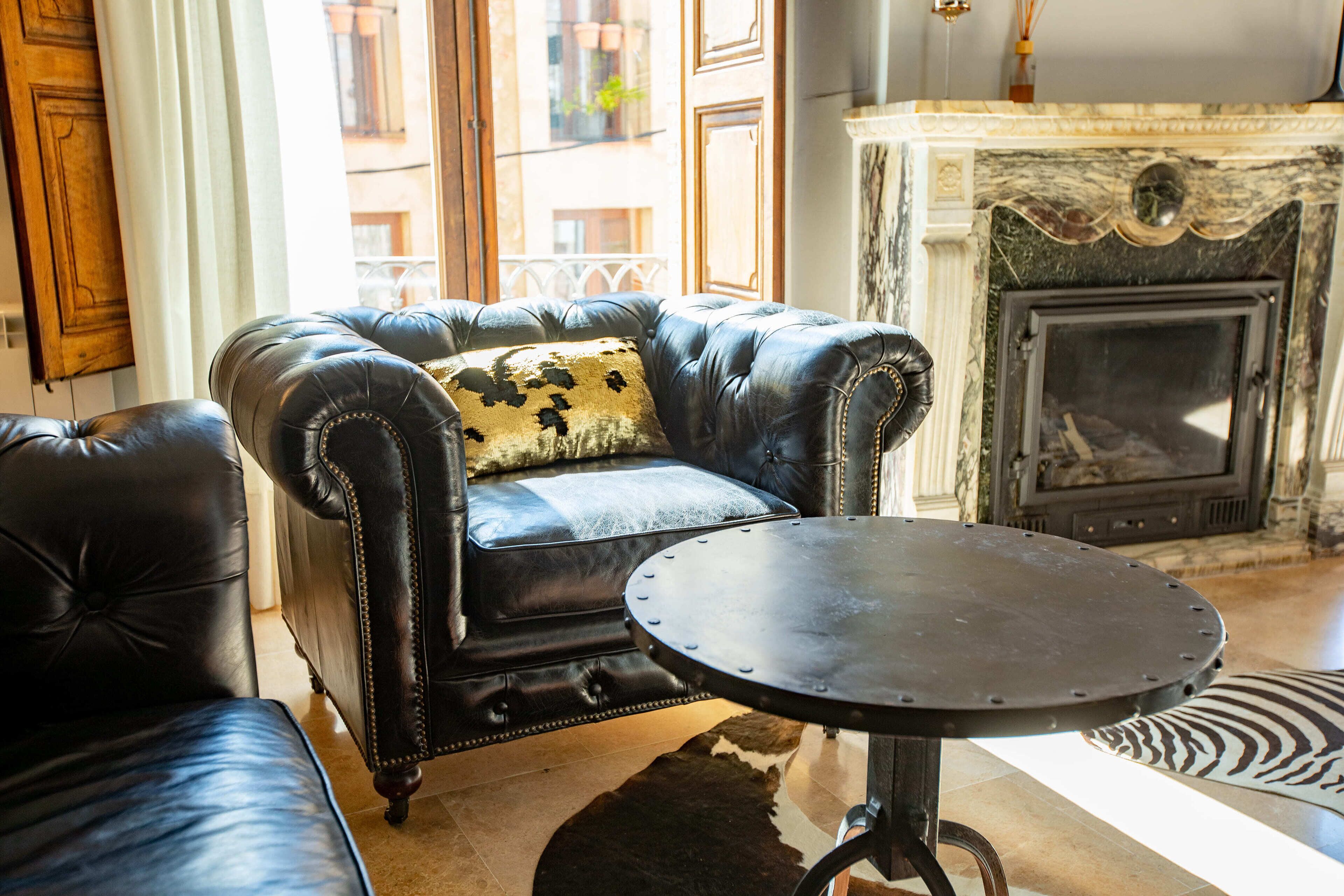 A leather sofa with a decorative pillow, a round metal table, and a fireplace in a well-lit room with large windows.