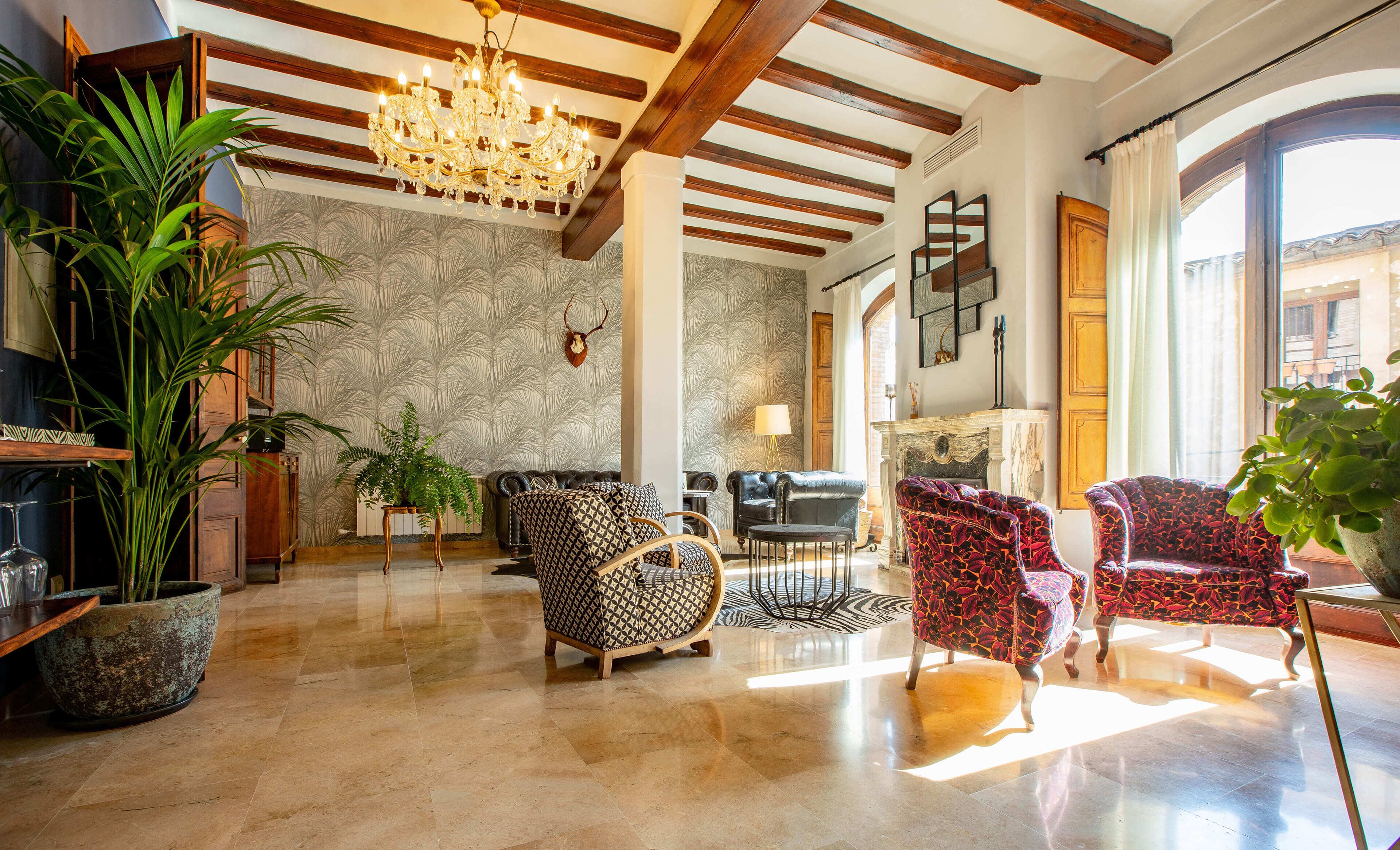 A spacious living area with a high ceiling, a chandelier, and a fireplace. There are several patterned armchairs and a large potted plant.